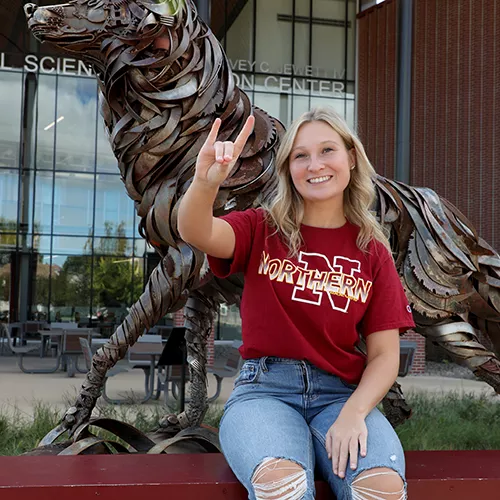 Female student sitting on outdoor bench with wolf sculpture behind her